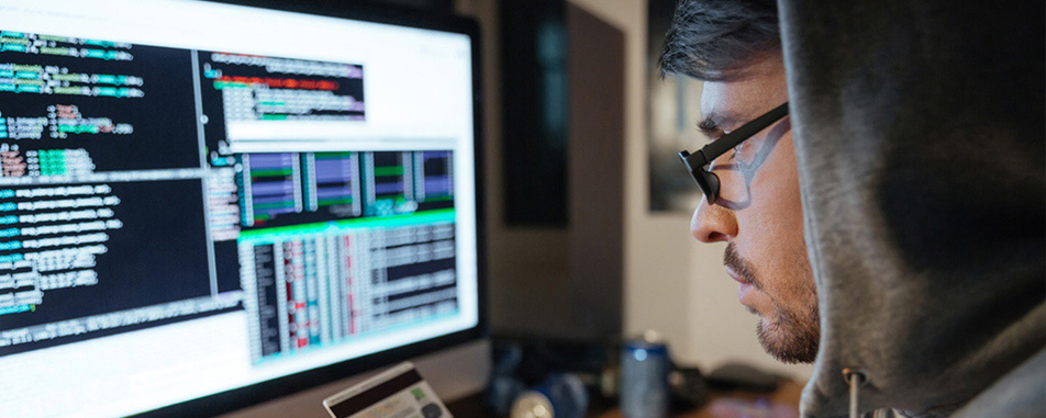A hacker in front of a computer monitor with screens of code visible, inputting credit card info. - www.quincymutual.com