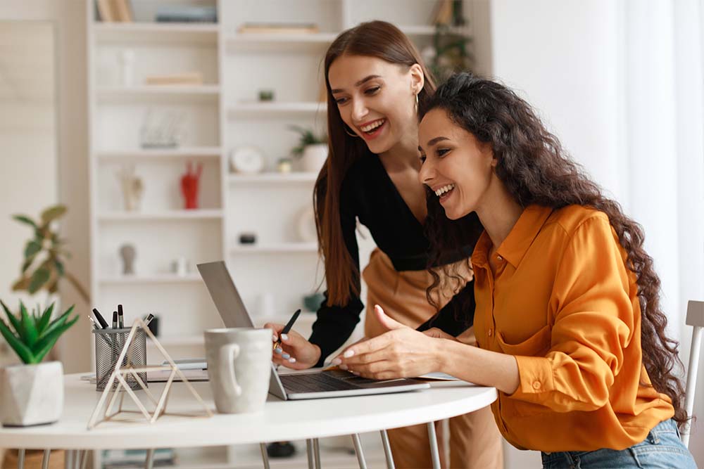 Two women laughing while collaborating at a desk with a laptop in a bright, home-office setting - https://www.quincymutual.com/index.htm