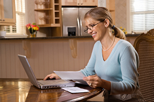 Woman at kitchen table working on her laptop with documents in her hand - https://www.quincymutual.com/