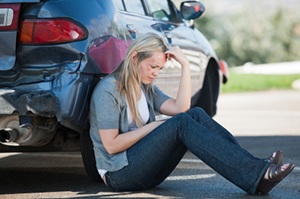 Upset woman sitting on the road with her back leaning against her car, which has a dented rear bumper. - https://www.quincymutual.com/